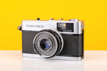 Load image into Gallery viewer, Olympus Trip 35 vintage 35mm film camera with Zuiko 40mm f2.8 lens, silver and black in color, against a yellow background.