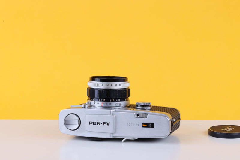 A vintage PEN-F-V camera with a silver body and black lens, positioned on a white surface against a solid yellow background, with its lens cap resting to the right.