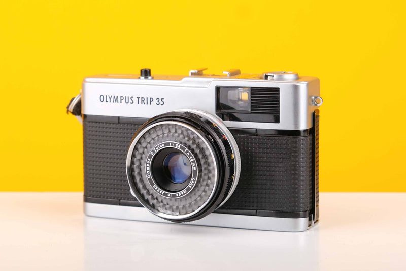 Olympus Trip 35 vintage 35mm film camera with Zuiko 40mm f2.8 lens, silver and black in color, against a yellow background.