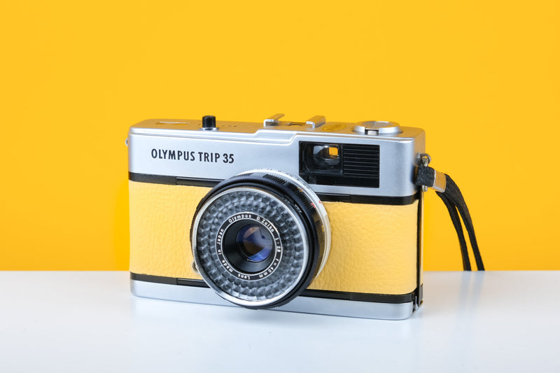 Olympus Trip 35 vintage film camera with Zuiko 40mm f2.8 lens, featuring a light yellow color, placed on a white surface against a yellow background.