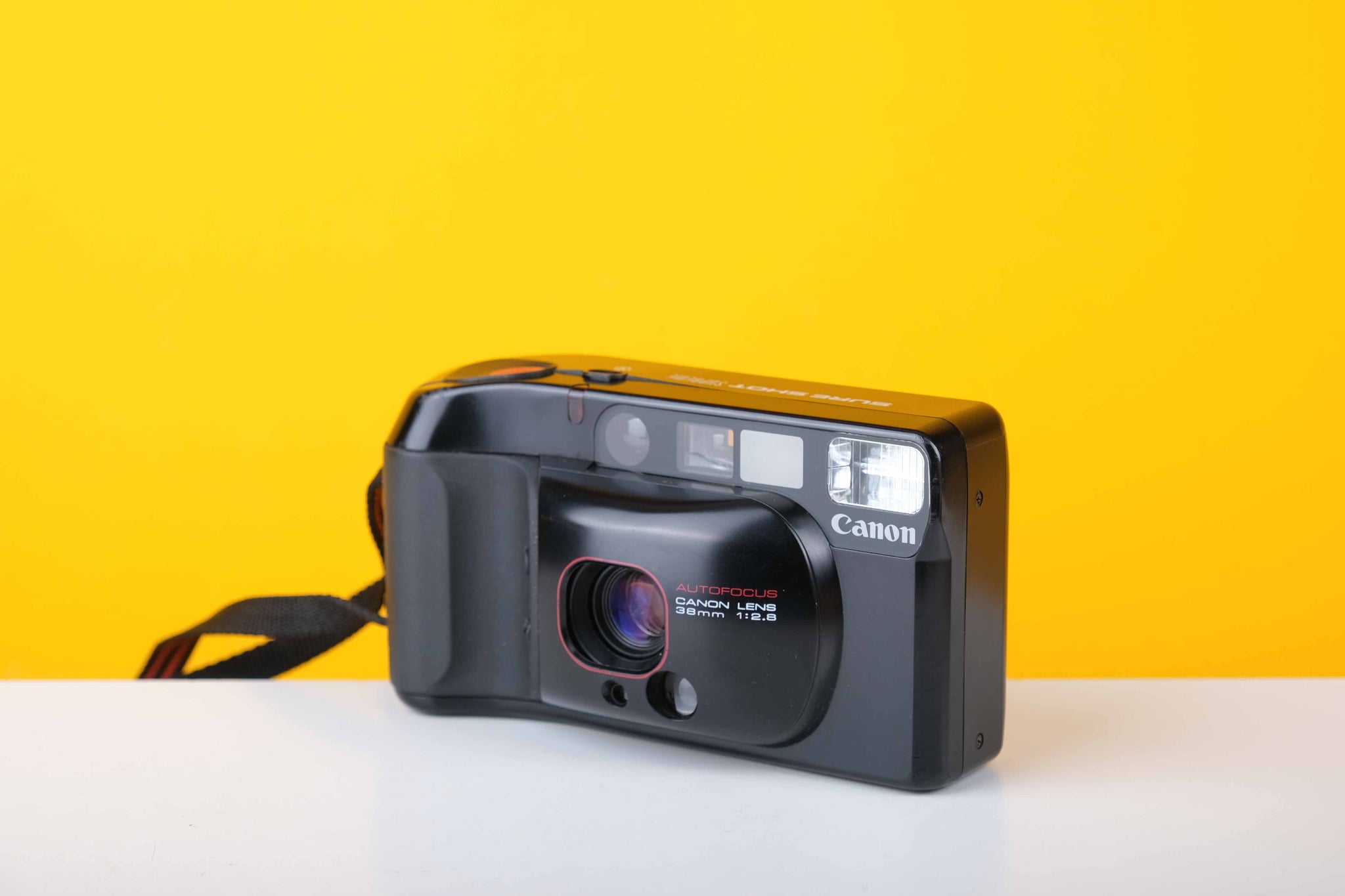 A Canon Sure Shot Supreme/Top Shot/Autoboy 3 vintage 35mm point and shoot film camera with a black casing, displayed against a yellow background.
