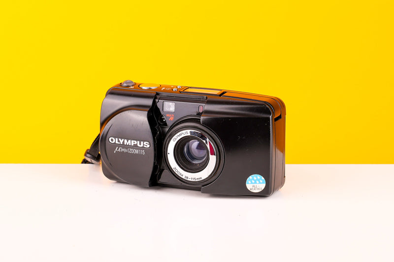 Olympus Mju Zoom 115 35mm Point and Shoot Film Camera with a zoom lens and built-in flash, displayed on a yellow and white background.