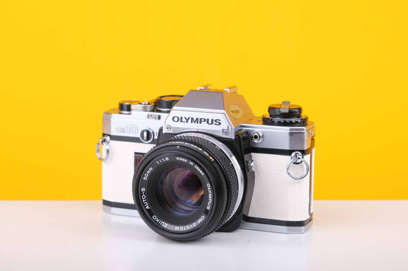 Olympus OM10 35mm Film Camera with a Olympus OM-System Zukio Auto-S 50mm f1.8 Lens on a white surface with a yellow background.