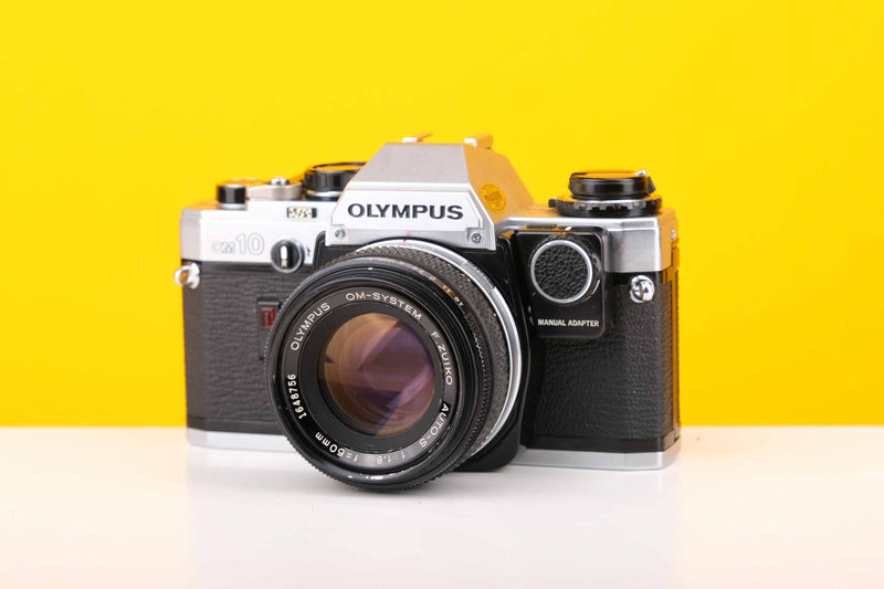 Olympus OM10 SLR 35mm Film Camera with Olympus Auto-S 50mm f1.8 MC Lens and Manual Adapter, displayed against a yellow background.