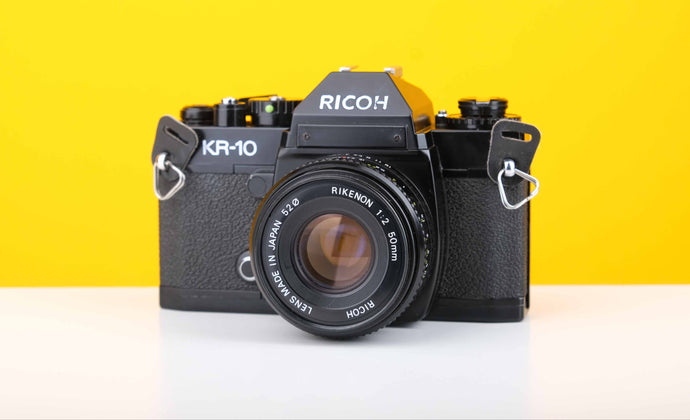 Ricoh KR-10 35mm SLR film camera with a Ricoh Rikenon 50mm f2 lens, displayed against a yellow background.