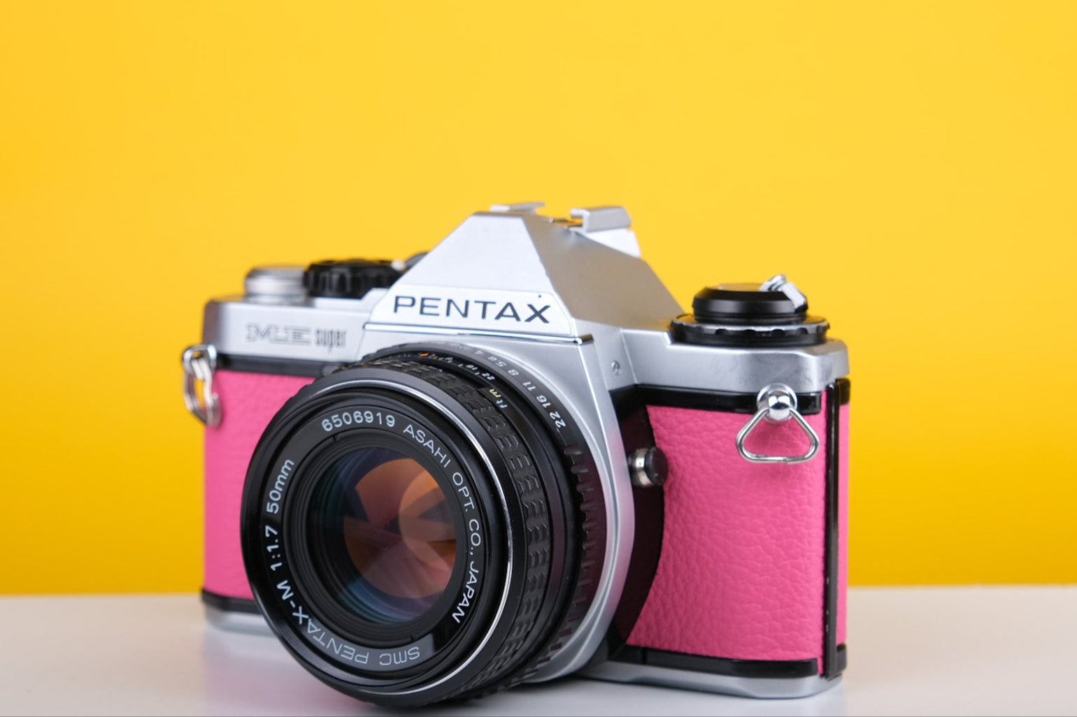 A pink Pentax ME Super 35mm SLR film camera with SMC Pentax-M 50mm f1.7 lens, placed on a white surface against a yellow background.